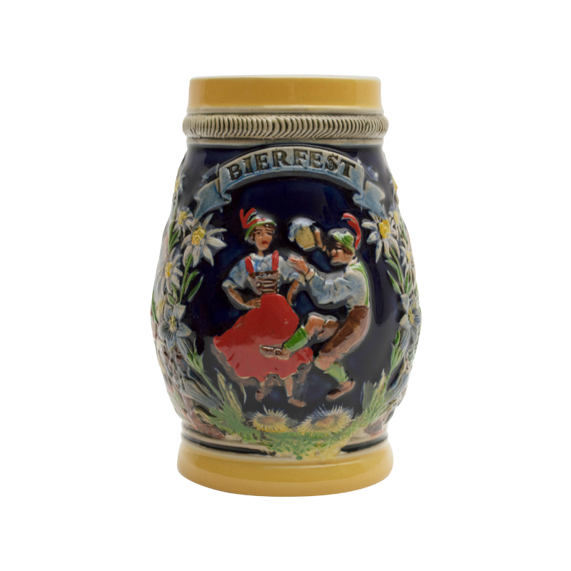 Decorative beer stein with a Bierfest theme, featuring embossed artwork of two individuals in traditional German attire, toasting with beer mugs. The central motif is framed by edelweiss flowers and greenery on a navy blue background, capped with a cream-colored rim with the word 'BIERFEST' prominently displayed.